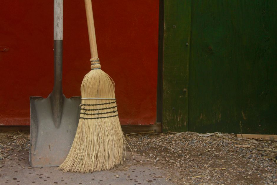 A garden spade and a straw broom leaning side by side against a shed door, ready for a clear-out